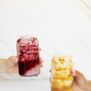 Two hands holding colorful iced drinks in clear glass jars against a bright backdrop.